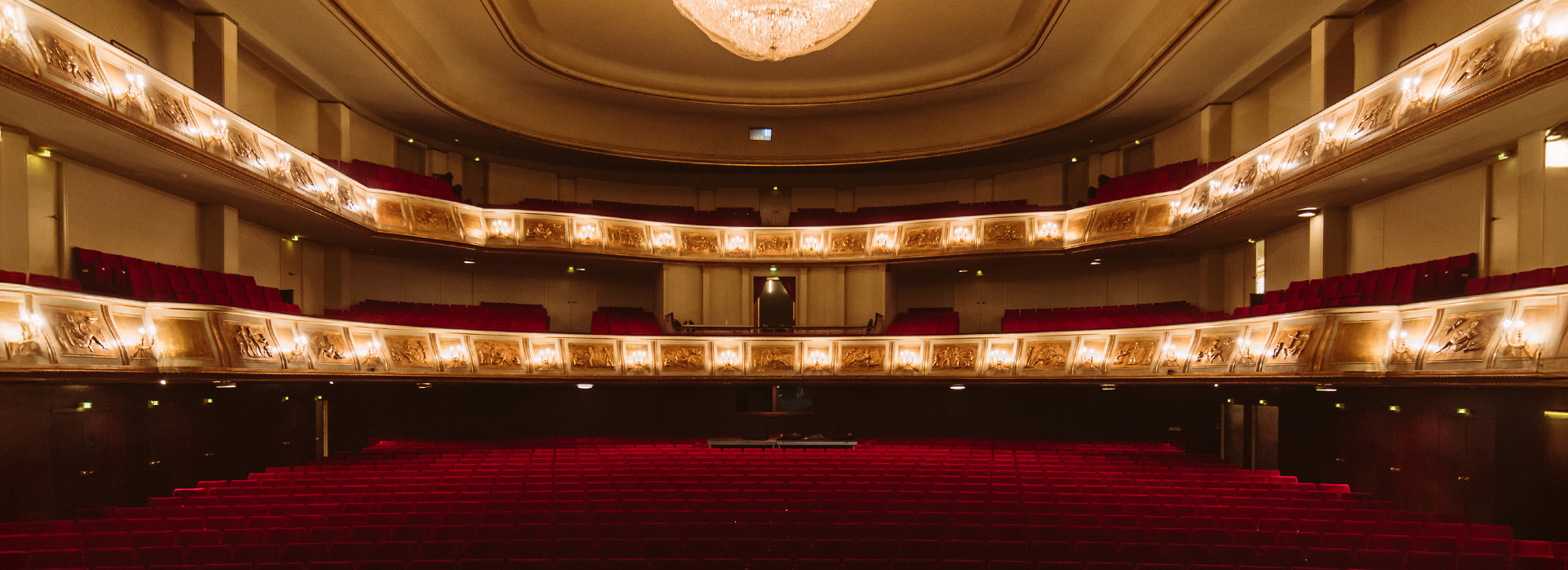 Blick aus dem Zuschauerraum auf die leeren roten Sitzreihen eines prunkvollen Theatersaals mit drei Rängen und einem großen Kronleuchter.