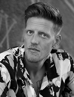 Porträtfoto von Edy Edwards mit Kurzhaarfrisur und markantem Blick, der ein Hemd mit künstlerischem Muster trägt. Das Bild ist in Schwarz-Weiß gehalten.