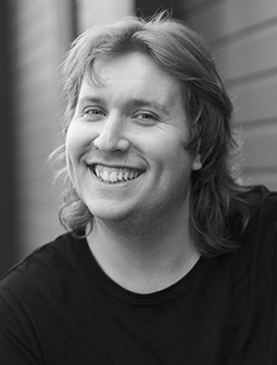 Porträtfoto von Linus Hencke mit mittellangen Haaren und einem breiten Lächeln. Er trägt ein dunkles T-Shirt. Das Bild ist in Schwarz-Weiß aufgenommen.