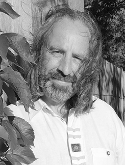 Porträtfoto von Dan Chamandy mit längeren Haaren und Bart, in einem hellen Hemd mit Natur im Hintergrund, Schwarz-Weiß