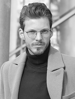 Porträtaufnahme von Niklas Schinke mit Brille und Mantel, vor urbanem Hintergrund, Schwarz-Weiß
