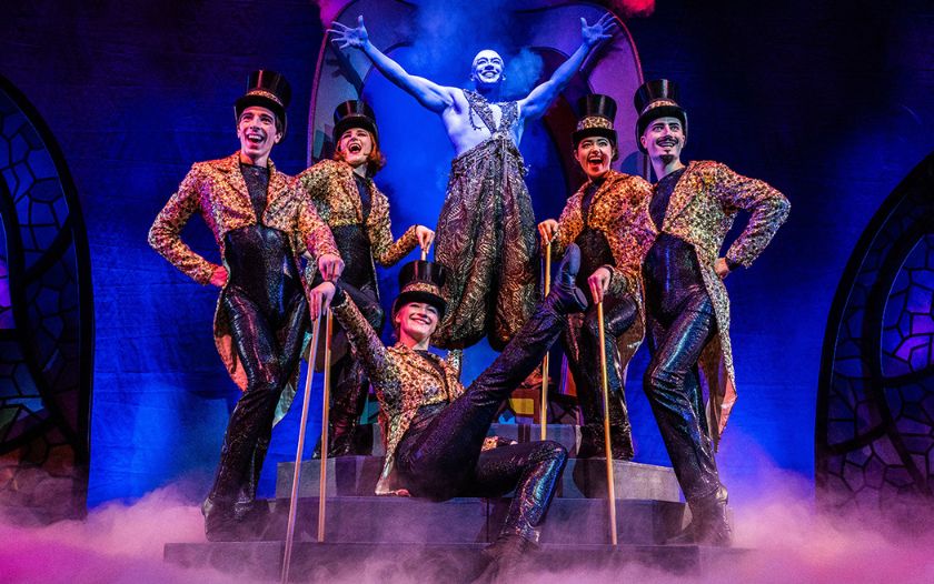 Der blaue Lampengeist steht triumphierend in der Mitte, umgeben von Tänzerinnen in Glitzerkostümen und Zylindern.