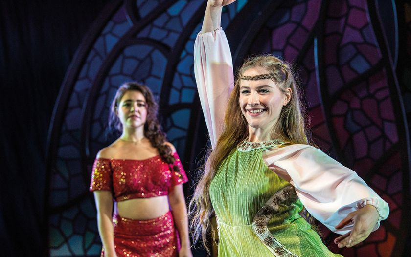 Eine Darstellerin in grünem Kleid tanzt freudestrahlend, im Hintergrund steht eine weitere Darstellerin in rotem Kostüm.