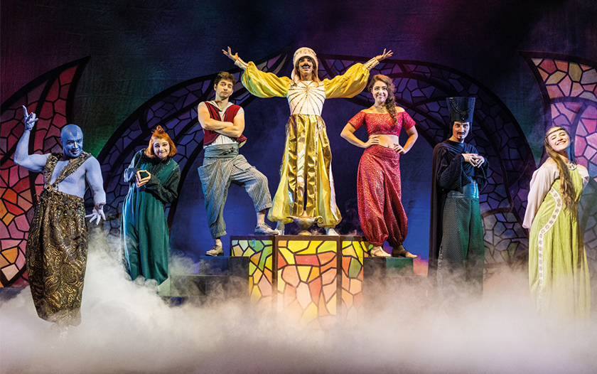 Das Ensemble von Aladin – das Musical von Theater Liberi steht auf der Bühne im Bühnenlicht und Nebel. In der Mitte der Bühne steht Aladin in goldener Kleidung erhoben, umgeben von weiteren bunten Charakteren, darunter ein blauer Dschinn, eine Frau in grünem Gewand und eine rotgekleidete Figur.