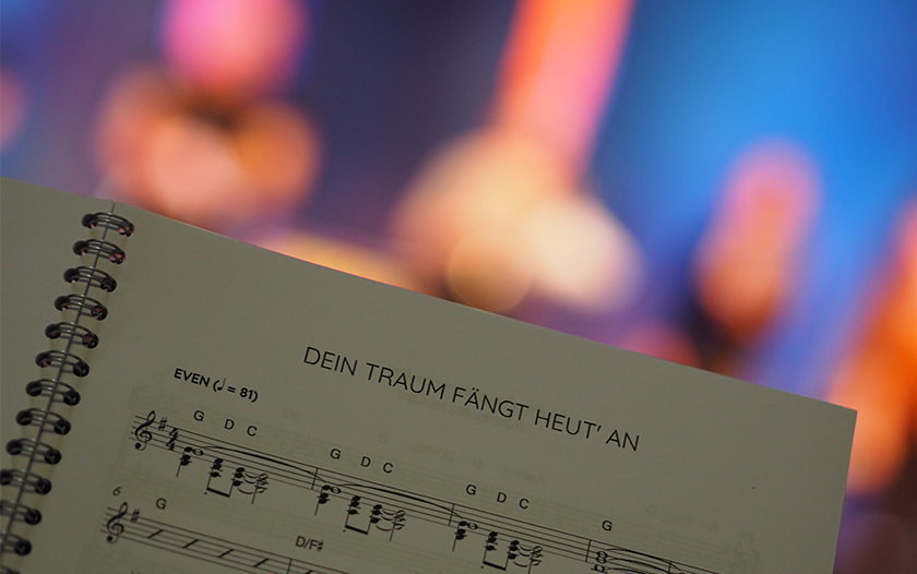 Nahaufnahme eines Notenblatts mit Titel 'Dein Traum fängt heut’ an' vor verschwommenem Bühnenlicht