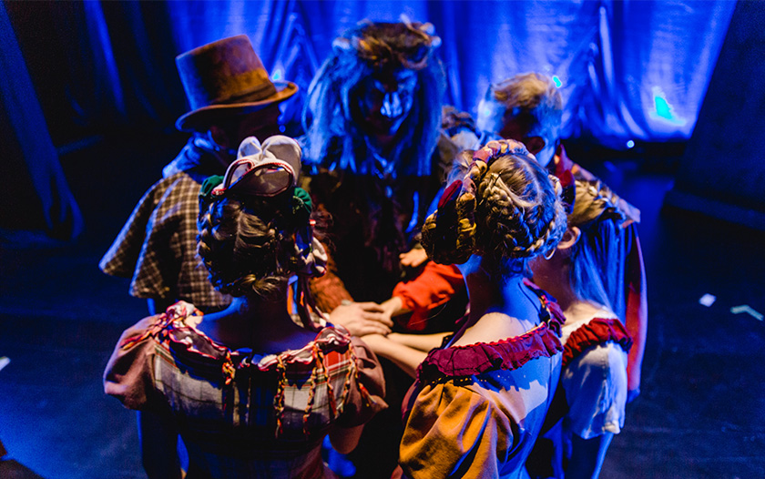 Darstellerinnen und das Biest stehen im Kreis hinter der Bühne, ihre Hände vereint in der Mitte, Bühnenlicht im Hintergrund.