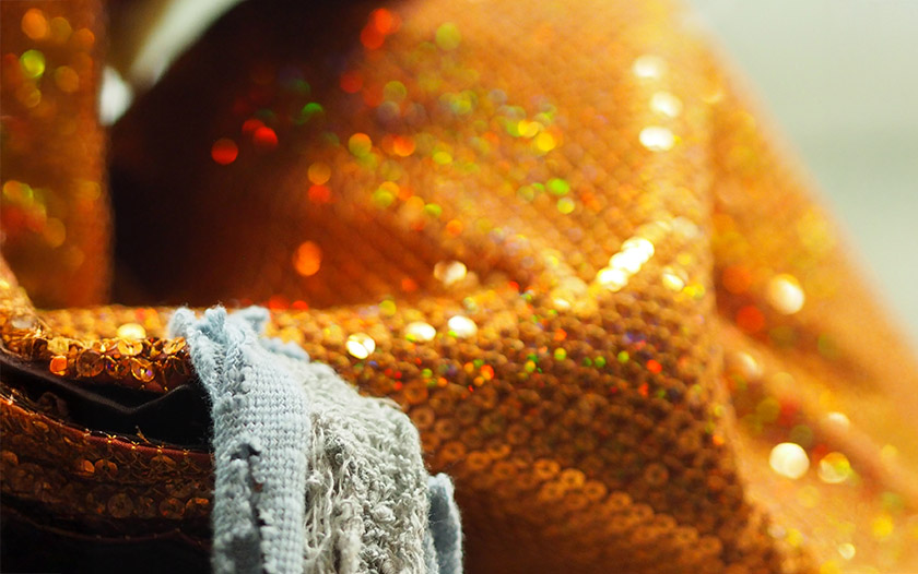 Ein glitzernder orangefarbener Stoff mit irisierenden Pailletten liegt in Falten, im Vordergrund ist ein ausgefranstes blaues Stoffstück zu sehen. Die intensive Farbigkeit und Textur zeigen einen markanten Bühnenstoff.