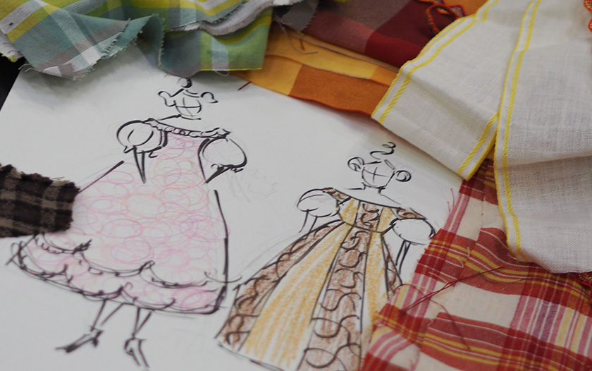 Zwei gezeichnete weibliche Figuren in historischen Kleidern auf einem Skizzenpapier, umgeben von bunten Stoffproben in Karomustern und warmen Farbtönen. Die Entwürfe zeigen fantasievolle Bühnenkleider mit Puffärmeln und verzierten Stoffbahnen.