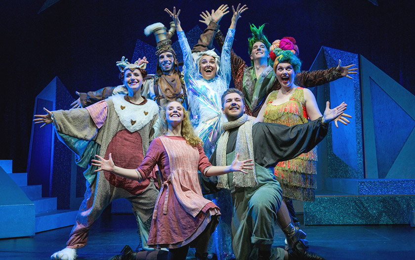 Sieben Darsteller in bunten Kostümen, darunter Schneekönigin, Gerda, Schlawuzis und weitere Figuren, posieren fröhlich auf der Bühne.