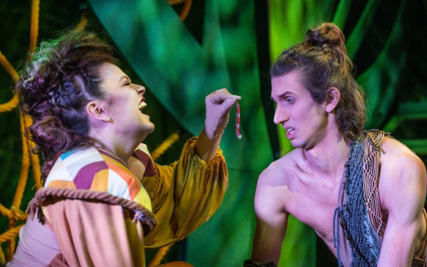 Eine Frau in bunter Dschungelkleidung lacht und zeigt Tarzan einen Wurm während er mit skeptischem Blick auf das Armband schaut.