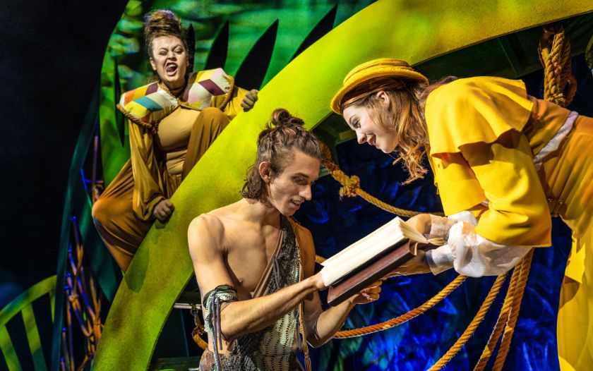 Jane, in gelbem Kleid, zeigt dem neugierigen Tarzan ein aufgeschlagenes Buch, während ein Affenwesen im Hintergrund erstaunt schaut.