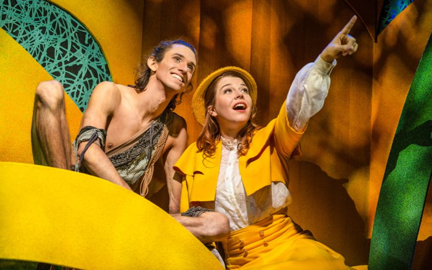 Szene aus dem Musical 'Tarzan – das Musical': Jane im gelben Kostüm zeigt mit ausgestrecktem Arm auf etwas in der Ferne, während Tarzan lächelnd neben ihr sitzt.