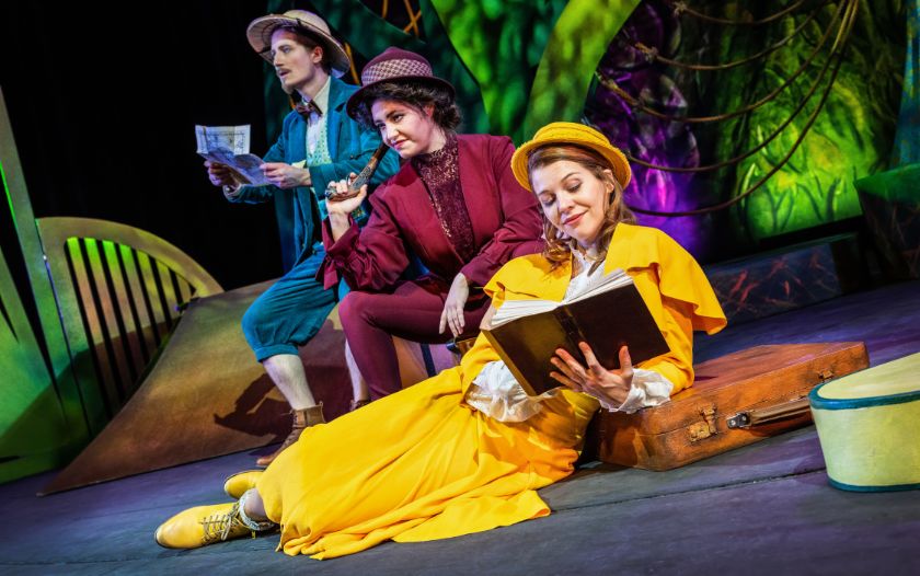 Jane liegt in einem gelben Kleid auf einem Koffer und liest ein Buch, während zwei weitere Figuren – einer mit Pistole – hinter ihr stehen.