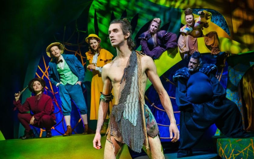 Tarzan steht mit aufrechter Haltung auf der Bühne, umgeben von Jane, Tee und weiteren Charakteren aus dem Musical.