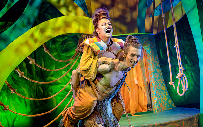 Tarzan mit nacktem Oberkörper trägt lachend eine Frau in gelbem Kostüm auf dem Rücken, beide in farbenfroher Dschungelkulisse.