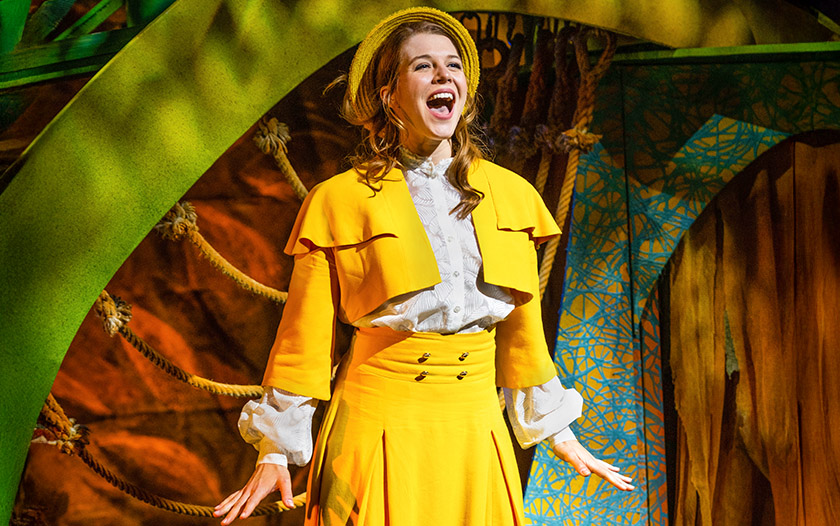 Frau im gelben Kostüm mit Hut singt strahlend vor einer grünen und orangen Dschungelkulisse.