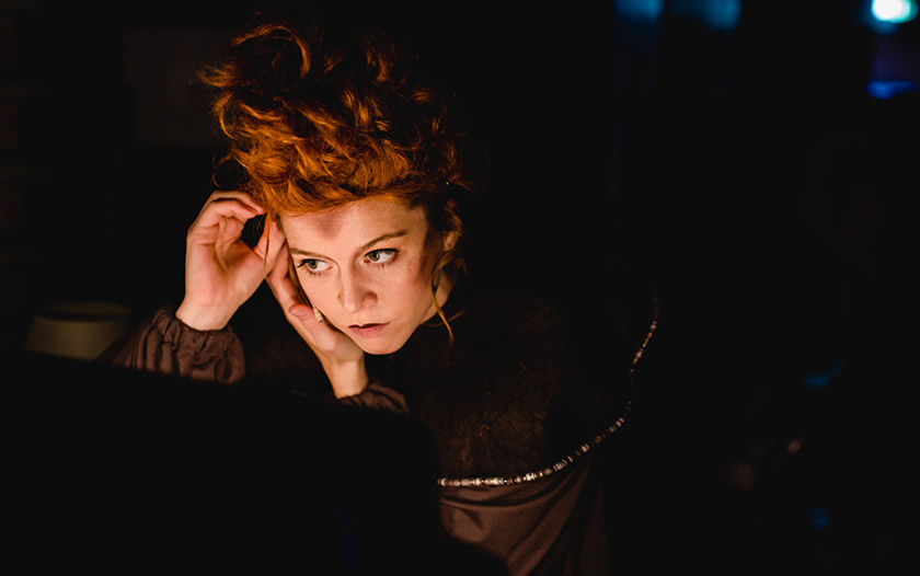 Darstellerin mit rotem, hochgestecktem Haar und dunklem Kostüm schaut konzentriert in den Spiegel, während sie sich im schwachen Bühnenlicht schminkt.