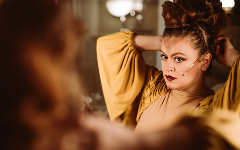 Schauspielerin im gelben Kostüm steht vor einem Spiegel und richtet mit beiden Händen ihre hochgesteckte Frisur, ein Mikrofon ist am Gesicht befestigt.