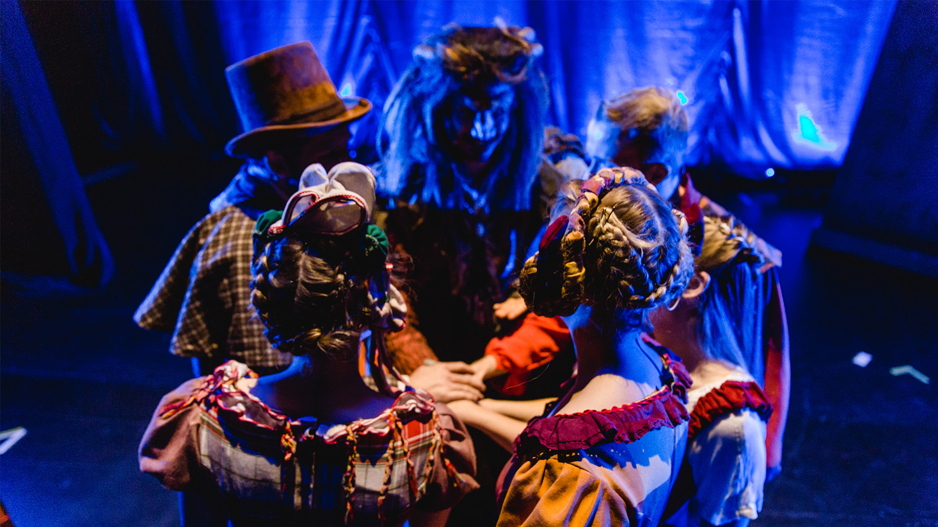 Ensemble von "Die Schöne und das Biest - das Musical" in bunten Kostümen steht im Kreis hinter der Bühne mit den Händen übereinander – Moment der Konzentration vor dem Auftritt bei Theater Liberi.