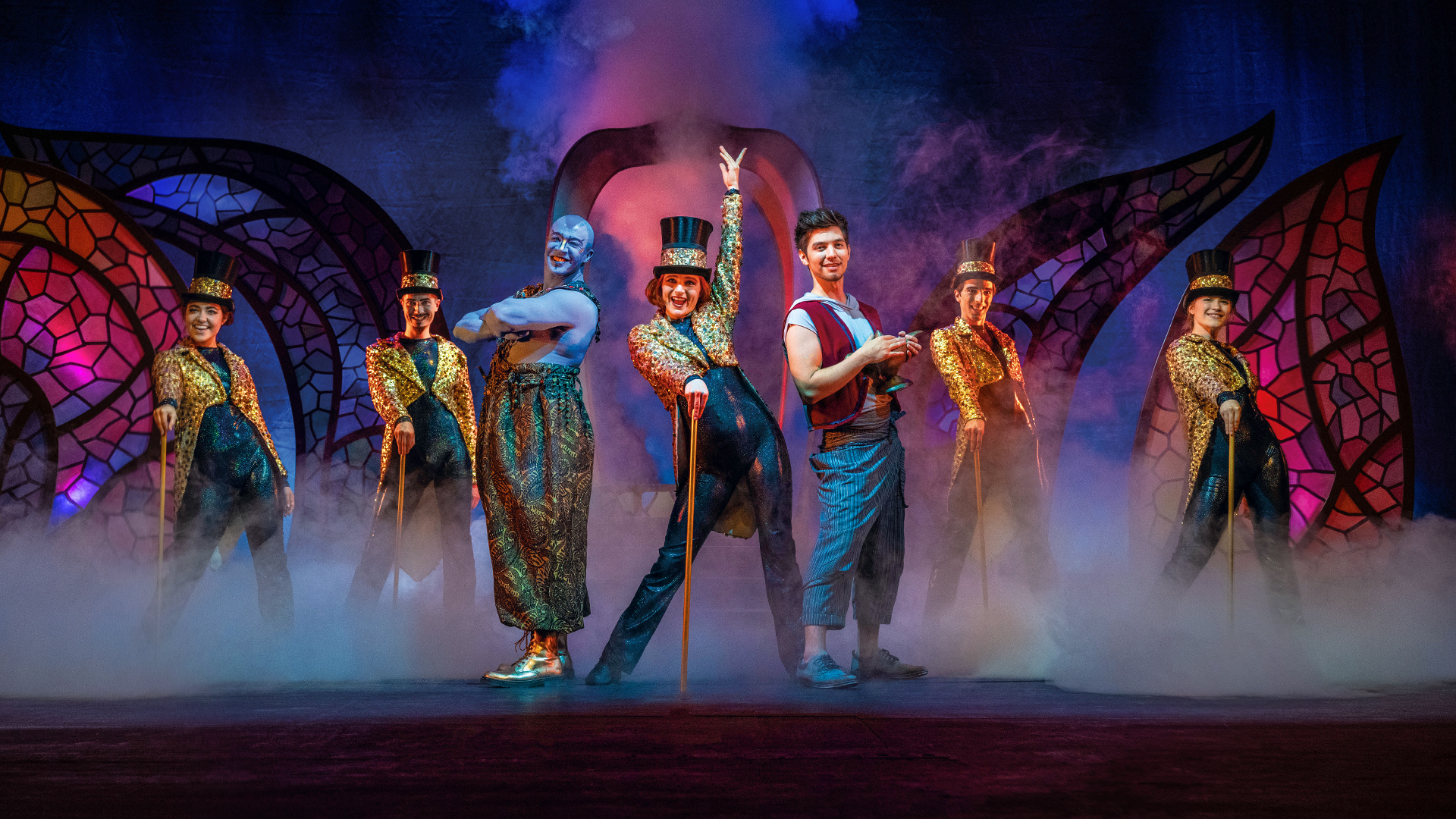 Ensemble von Theater Liberi in fantasievollen Kostümen auf der Bühne: Aladin, Dschinni und Tänzer:innen in Glitzeroutfits mit Zylindern, umgeben von Bühnennebel und orientalischem Bühnenbild.
