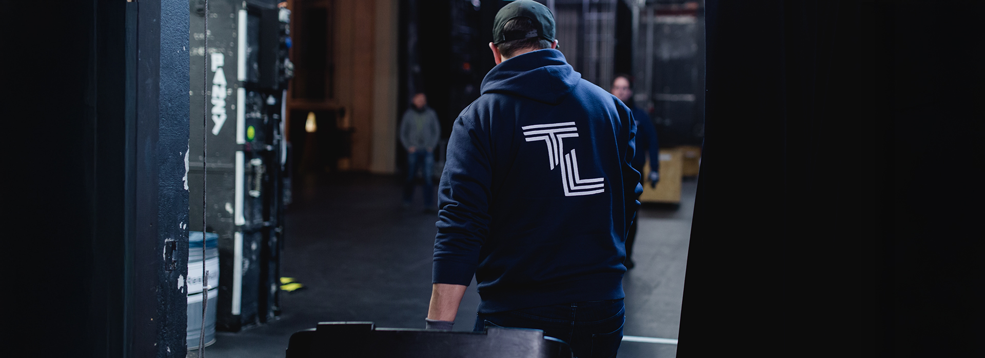 Blick hinter die Bühne: Ein Techniker mit TL-Hoodie schiebt einen Transportkoffer durch den Bühnenbereich während des Aufbaus.