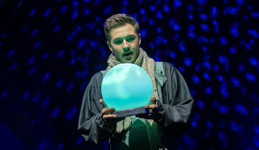Schauspieler auf der Bühne hält eine leuchtende, türkisfarbene Kristallkugel, während er in einem dramatisch beleuchteten Bühnenbild mit blauem Sternenlicht steht.