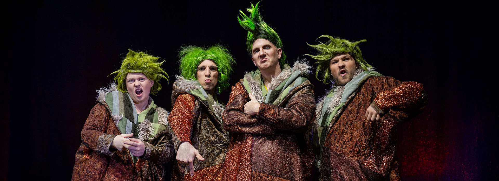 Vier Darsteller in glitzernden, braun-orangenen Schlawuzi-Kostümen mit wilden, grünen Perücken stehen auf der Bühne und machen freche Posen.