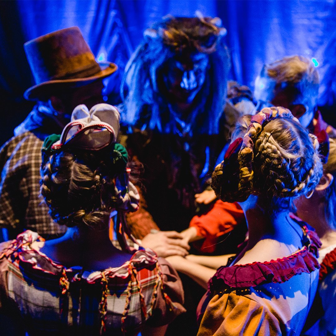 Ensemble von "Die Schöne und das Biest - das Musical" in bunten Kostümen steht im Kreis hinter der Bühne mit den Händen übereinander – Moment der Konzentration vor dem Auftritt bei Theater Liberi.