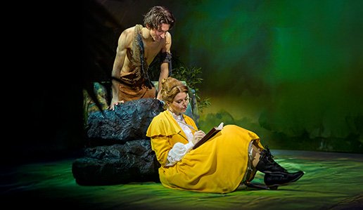 Eine Darsteller:in sitzt im Jane-Kostüm auf dem Boden und zeichnet in einem Buch. Ein Darsteller im Tarzan-Kostüm schaut ihr von oben über die Schulter.