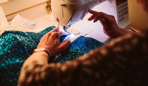 Kostümherstellung für Theater Liberi Nahaufnahme von Händen, die an einer Nähmaschine ein glitzerndes türkisfarbenes Kostüm nähen – im Hintergrund ein beleuchteter Schminkspiegel.