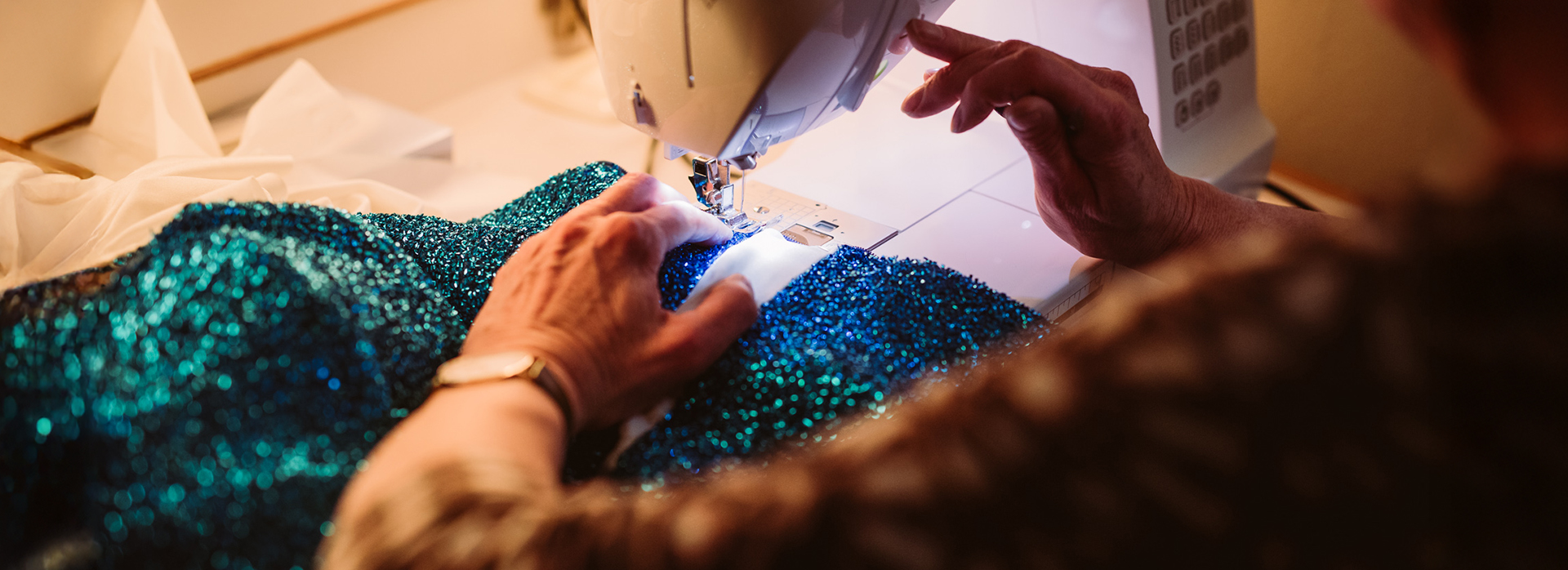 Nahaufnahme von Händen, die an einer Nähmaschine ein glitzerndes türkisfarbenes Kostüm nähen – im Hintergrund ein beleuchteter Schminkspiegel.