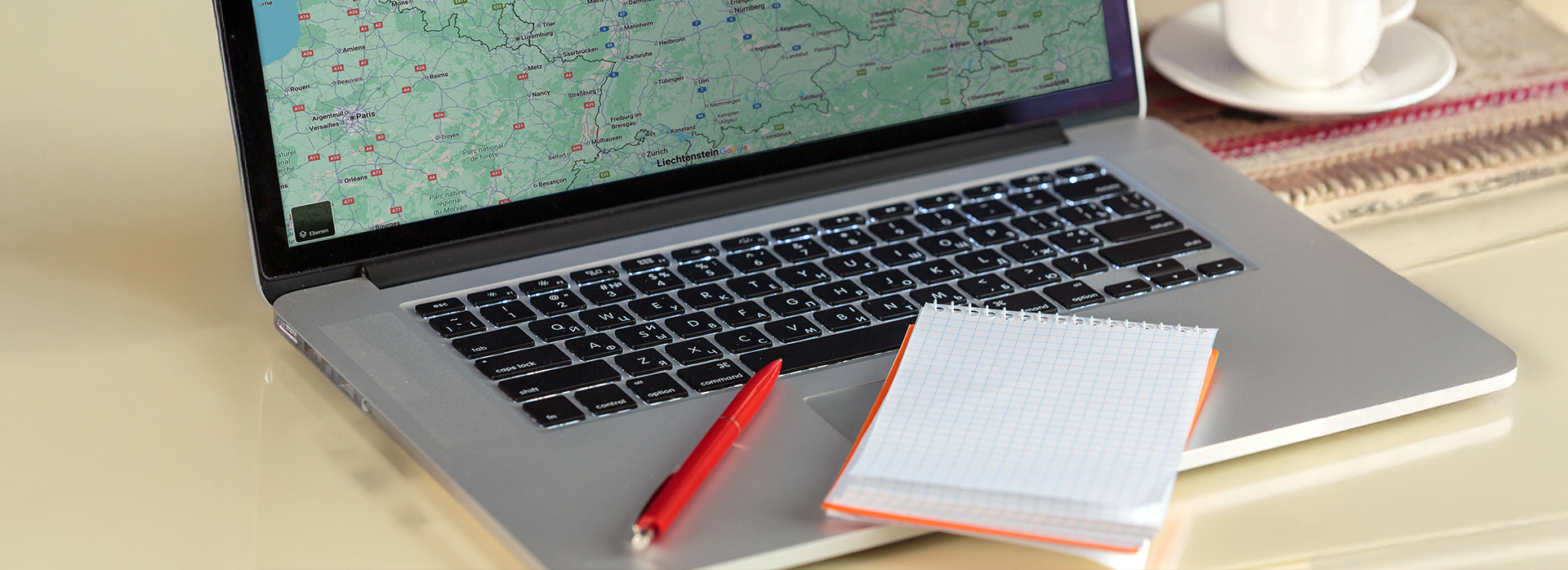 Laptop mit geöffneter Landkarte, daneben ein roter Stift, ein kariertes Notizbuch und ein Kaffee – Symbol für die Arbeit im Büro bei Theater Liberi.