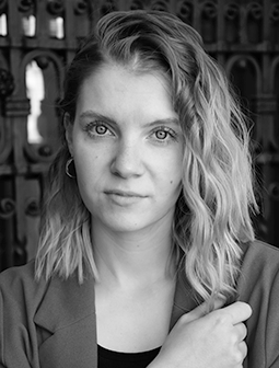 Schwarz-weißes Porträtfoto der Darstellerin Lisa-Marie Breithaupt aus dem Ensemble von Theater Liberi. Sie blickt ernst in die Kamera, trägt offene, leicht gewellte Haare und eine Jacke vor ornamentalem Hintergrund.