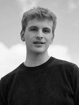 Portraitfoto von Moritz Ende in einem dunklen Pullover, stehend vor einem hellen Himmel, mit neutralem Gesichtsausdruck, monochrom