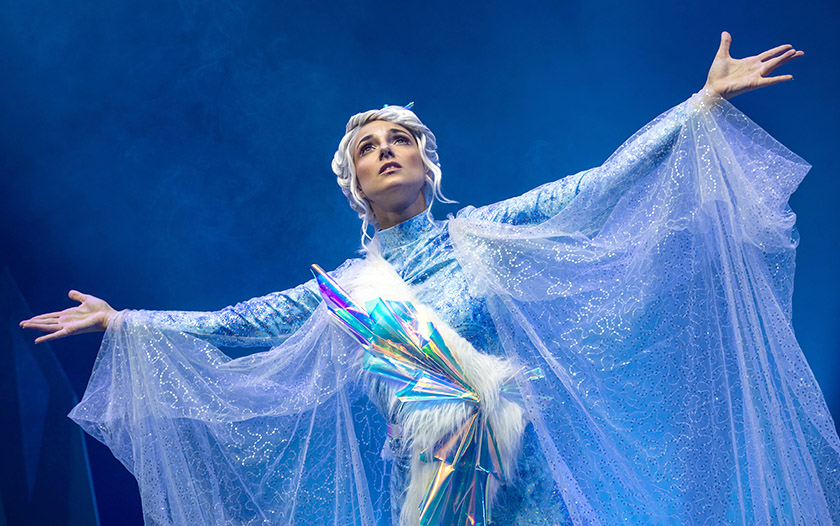 Darstellerin der Schneekönigin in glitzerndem, hellblauem Kostüm mit ausgebreiteten Armen vor blauem Bühnenhintergrund.