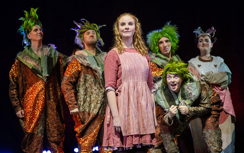 Junge Frau in rosa Kleid (Gerda) steht inmitten der Schlawuzis mit grünen Perücken und braun-orangenen Kostümen.