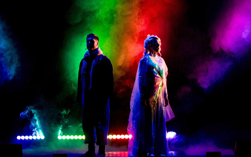 Zwei Darsteller, Kay und die Schneekönigin, stehen mit dem Rücken zueinander auf einer Bühne, umgeben von farbigem Licht in Grün, Rot, Lila und Blau.