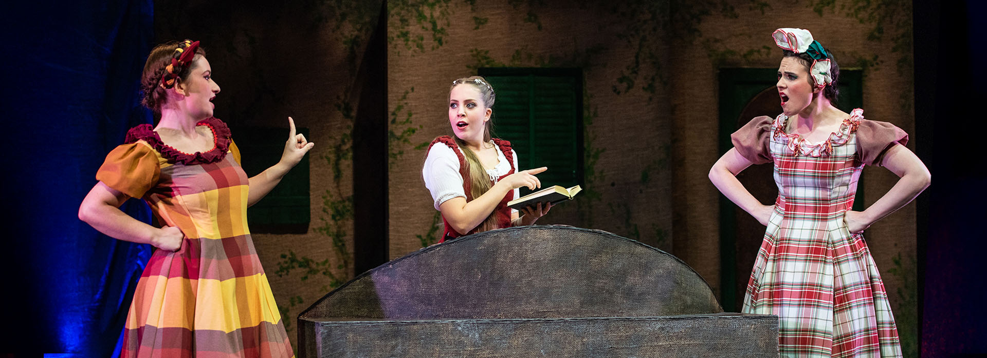 Drei Darstellerinnen in bunten Kostümen stehen um einen großen Brunnen; eine von ihnen hält ein Buch – es ist Belle.