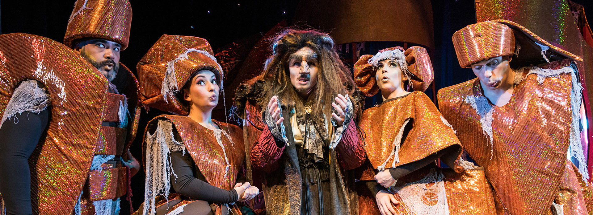 Eine Gruppe Darsteller in fantasievollen, kupferfarbenen Kostümen steht um das Biest mit zotteliger Mähne in der Mitte.