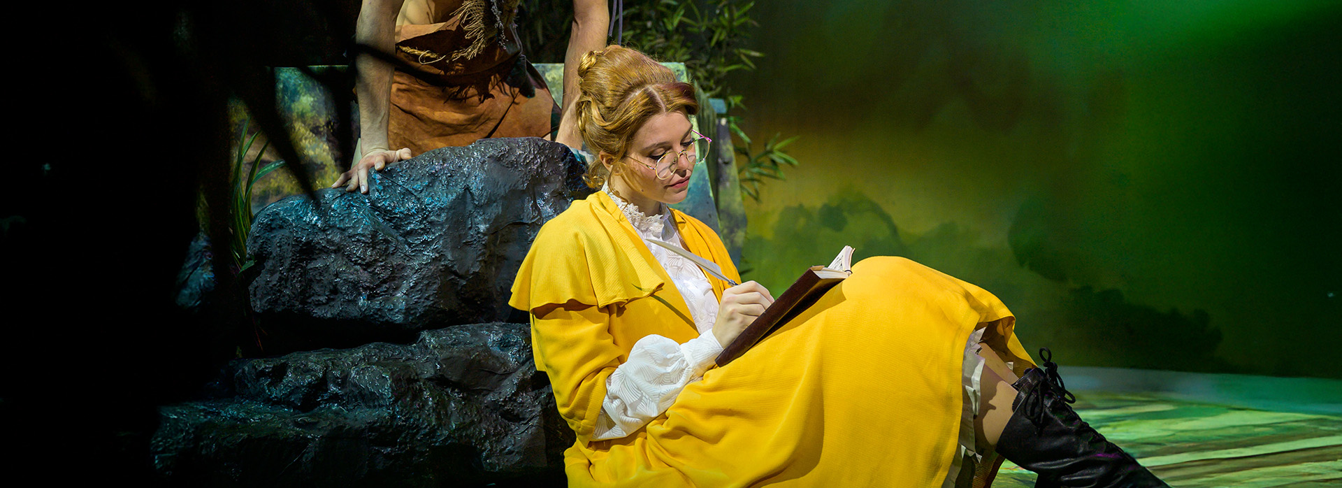 Eine Darsteller:in sitzt im Jane-Kostüm auf dem Boden und zeichnet in einem Buch. Ein Darsteller im Tarzan-Kostüm schaut ihr von oben über die Schulter.