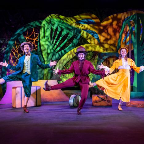 Drei Darsteller in bunten Kostümen tanzen vor lebendigem Bühnenbild.