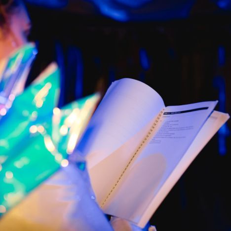 Eine Person hält ein aufgeschlagenes Skript in der Hand, während sie in einem Kostüm mit blaugrünen Spitzen auf den Auftritt wartet.