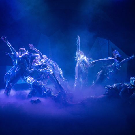 Tänzer:innen in reflektierenden silber-blauen Kostümen bewegen sich dramatisch im Bühnennebel.