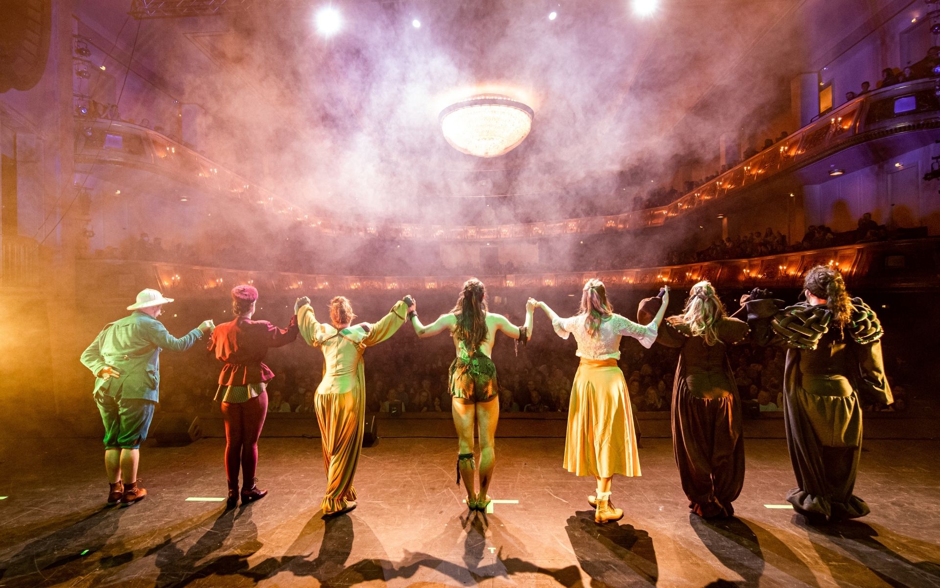  Ensemble steht Hand in Hand auf der Bühne vor Publikum im Theaterlicht.